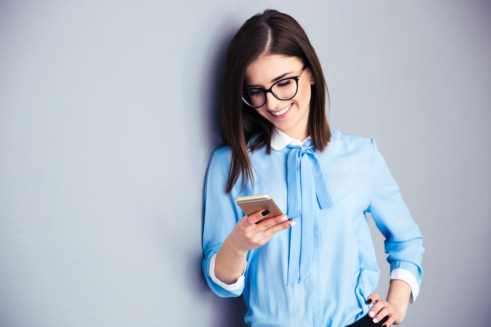Happy businesswoman using smartphone over gray background. Wearing in blue shirt and glasses.